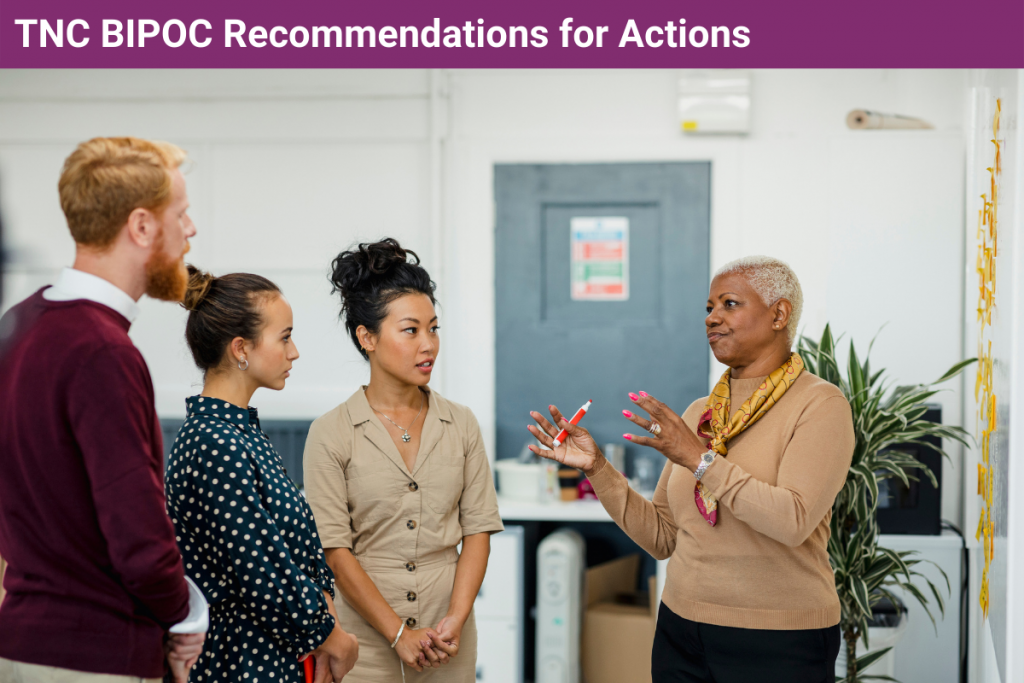 photo of a woman leading a conversation with three other people, all standing up in front of a wall with sticky notes on it